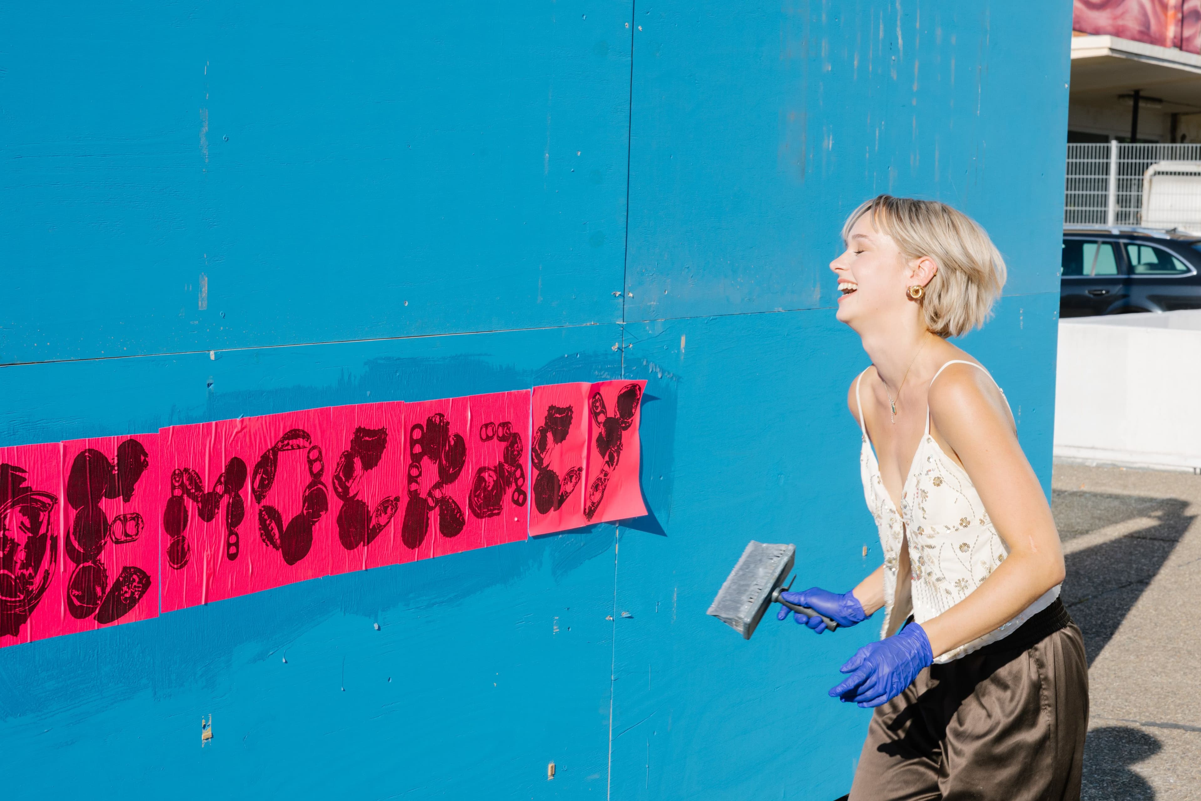 Eine Person kleistert ein Schild an eine blaue Wand und lacht.