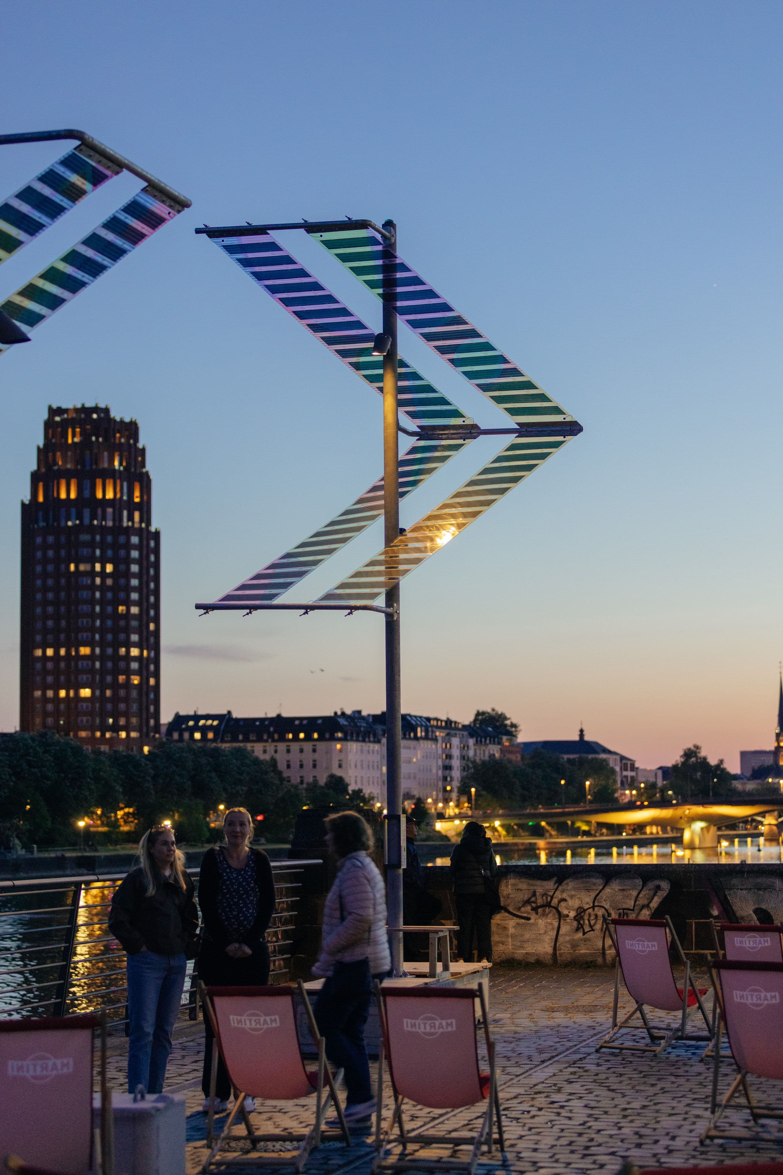 Zwei Menschen stehen unter einer dreieckigen Beleuchtung vor der Frankfurter Skyline im Nachthimmel.