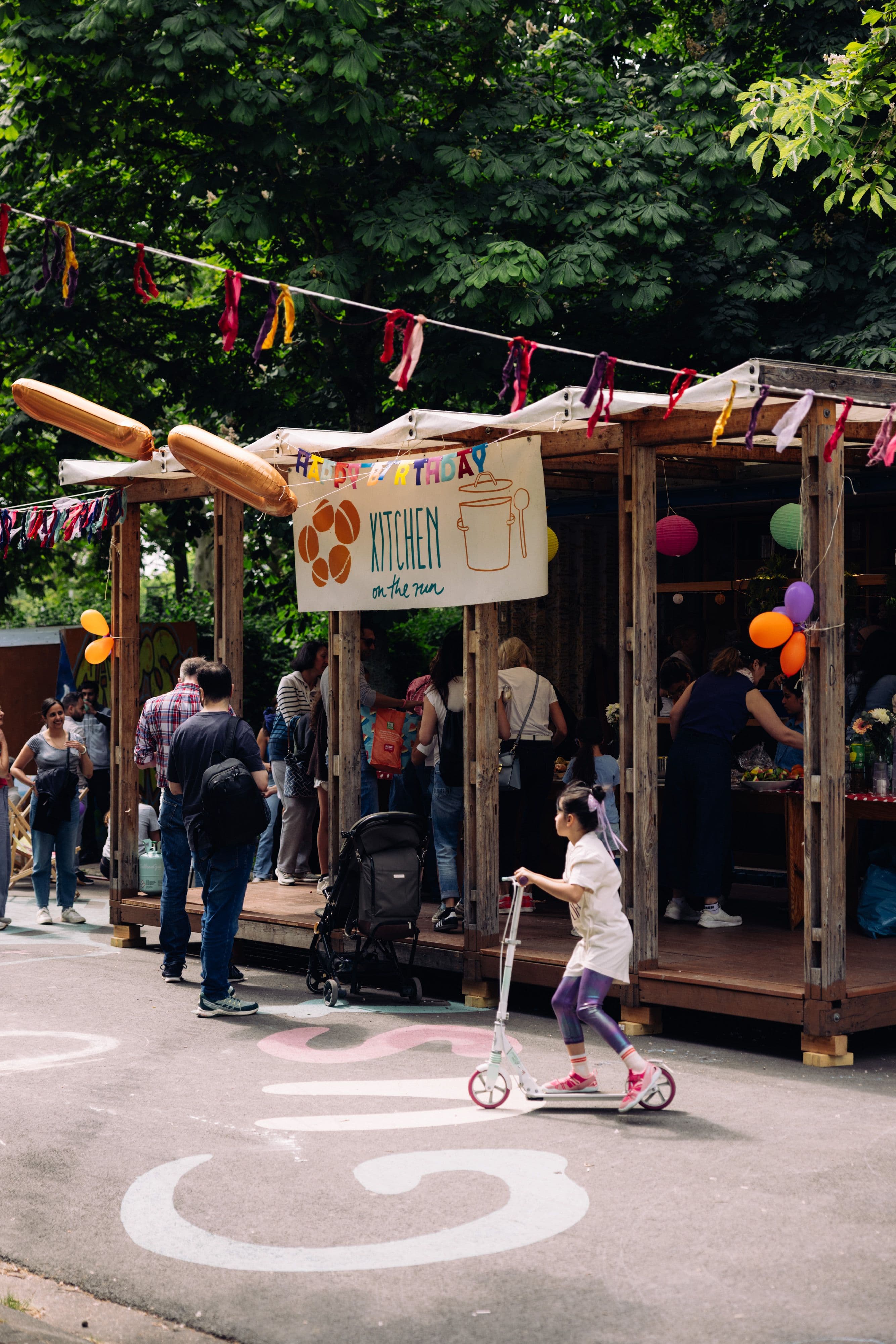 Mehrere Personen sind auf einer Art Straßenfest vor einem Container mit vielen Luftballons und Girlanden.
