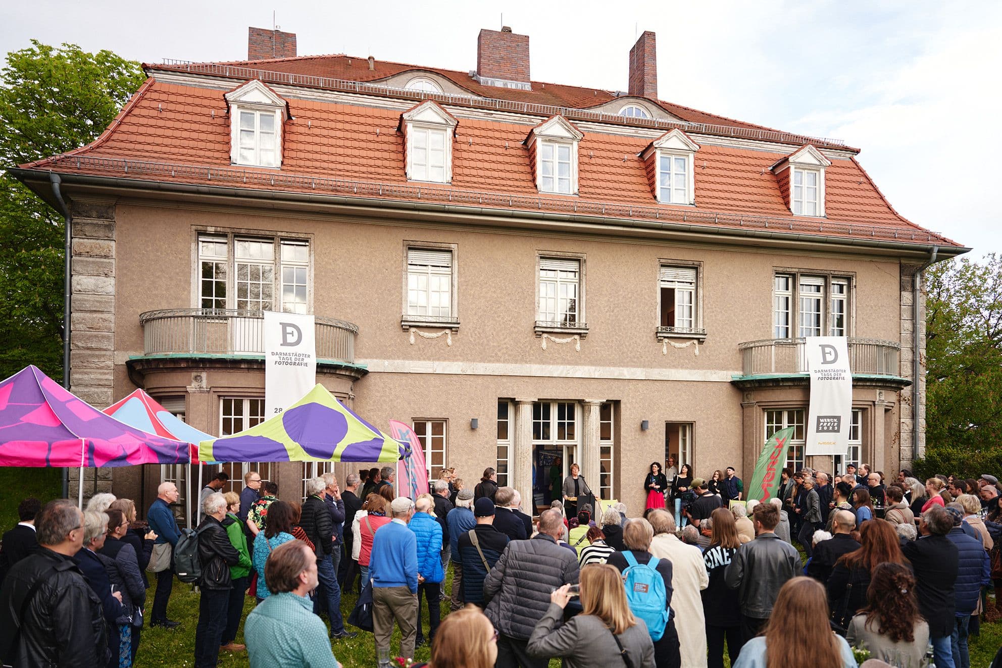 Vor einem Haus sind einige Leute versammelt, die einer Rede zuhören.