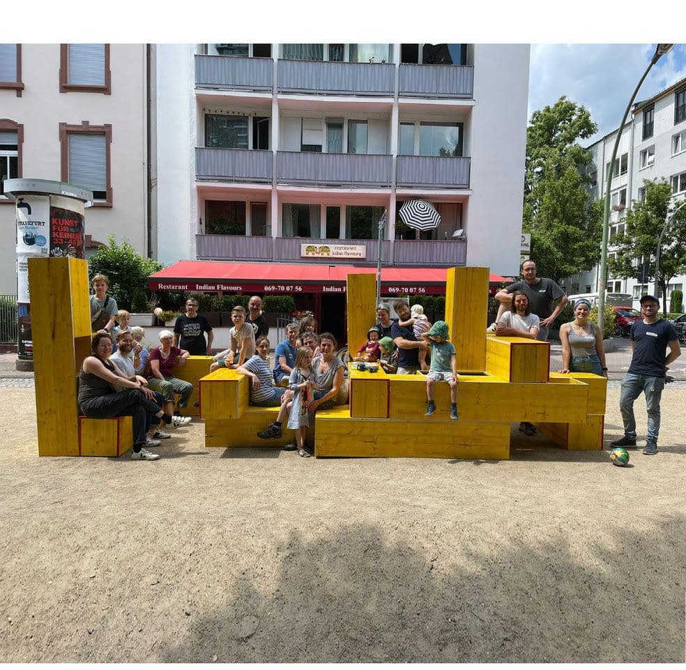 Eine Gruppe von Menschen sitzt auf gelben Holzklötzen auf der Straße.