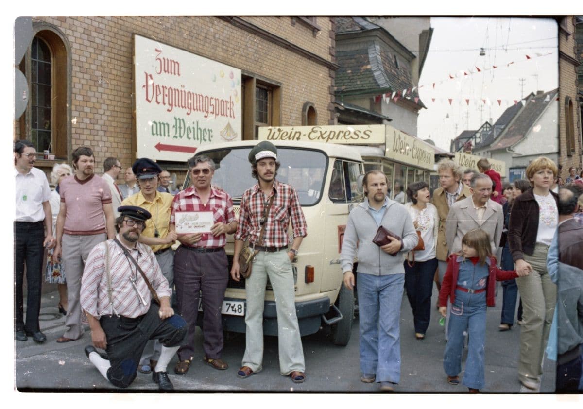 Vor einem Wagen mit der Aufschrift „Wein-Express” stehen viele Menschen auf der Straße.