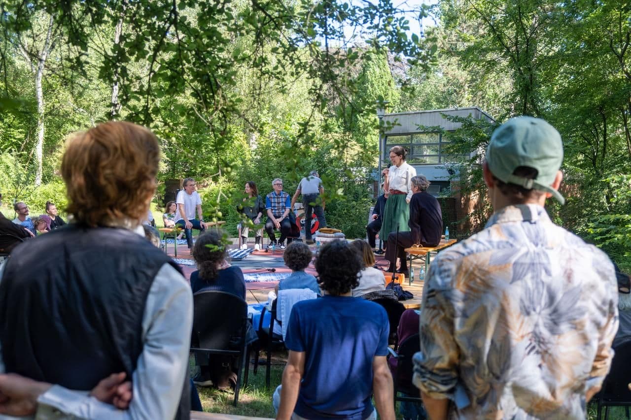 Menschen hören dem Vortrag einer Person auf einer Open-Air Bühne zu.