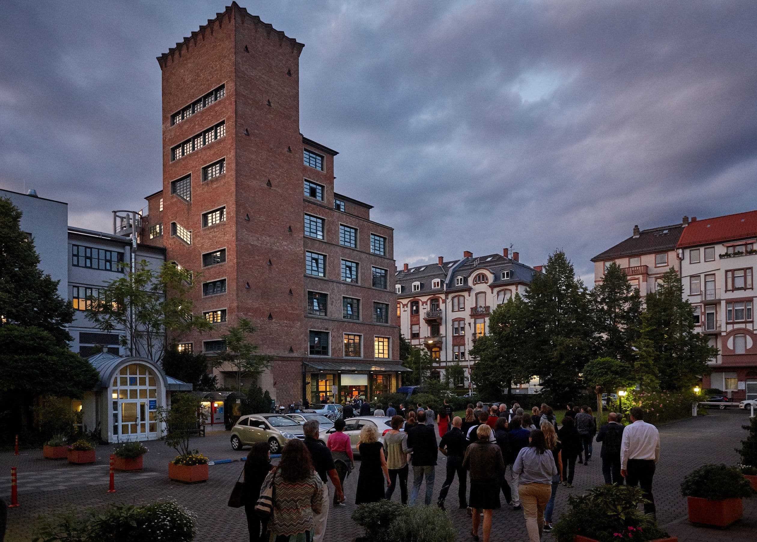 Der Mousonturm im Abendlicht: Ein hohes Gebäude aus roten Klinkersteinen mit vielen Fenstern. Davor stehen einige Menschen.