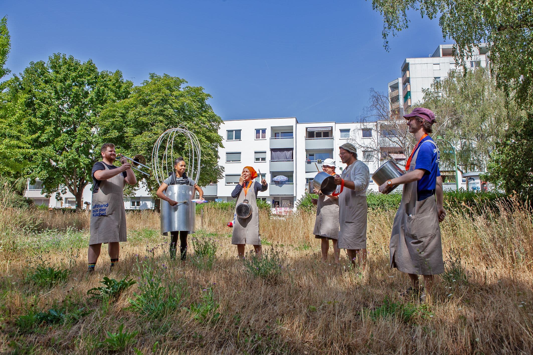 Mehrere Personen haben sich als Koch-Utensilien verkleidet und stehen auf einer Wiese vor einer Siedlung.