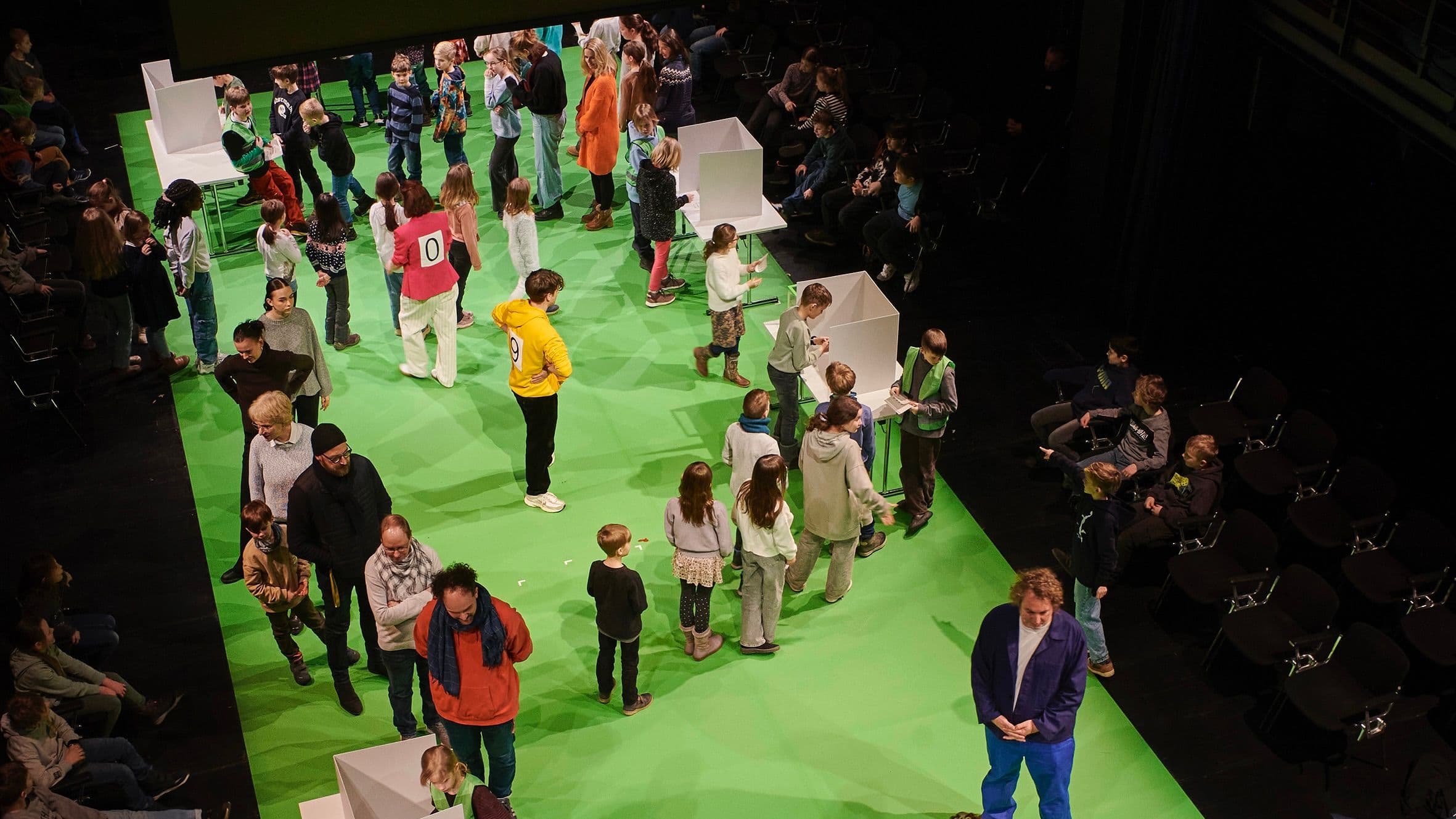 Viele Kinder und Erwachsene simulieren auf einer Bühne eine Wahlkabine