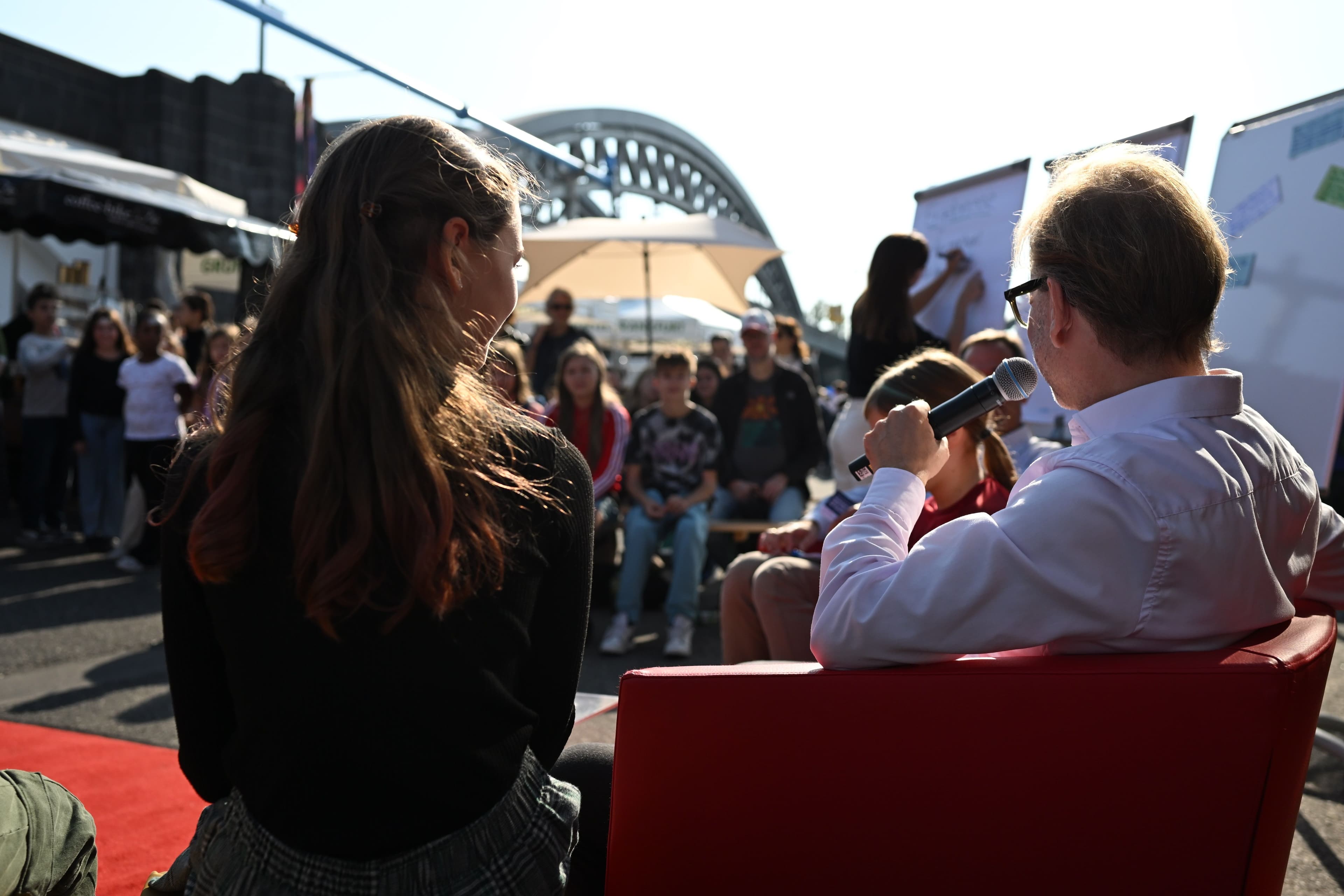 Von hinten sind ein Mädchen und ein Mann mit Mikrofon zu sehen, wie sie vor Kindern im Publikum draußen reden.