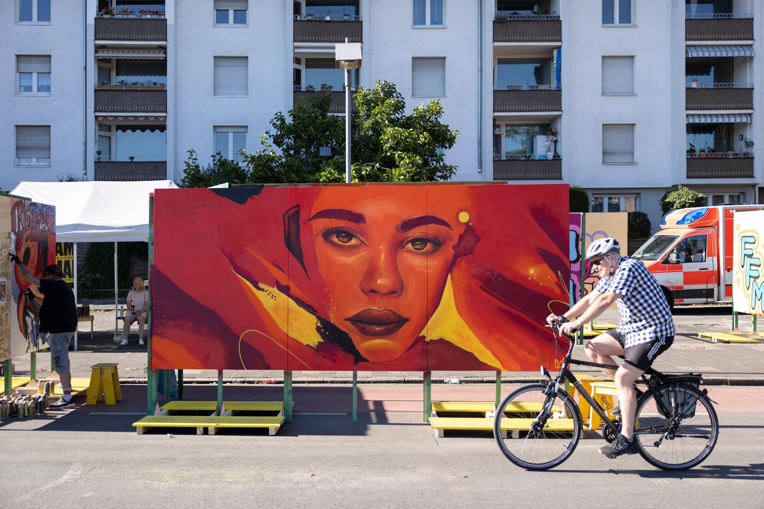 Auf einer Straße steht ein großes Gemälde, vor dem ein Fahrradfahrer vorbeifährt.