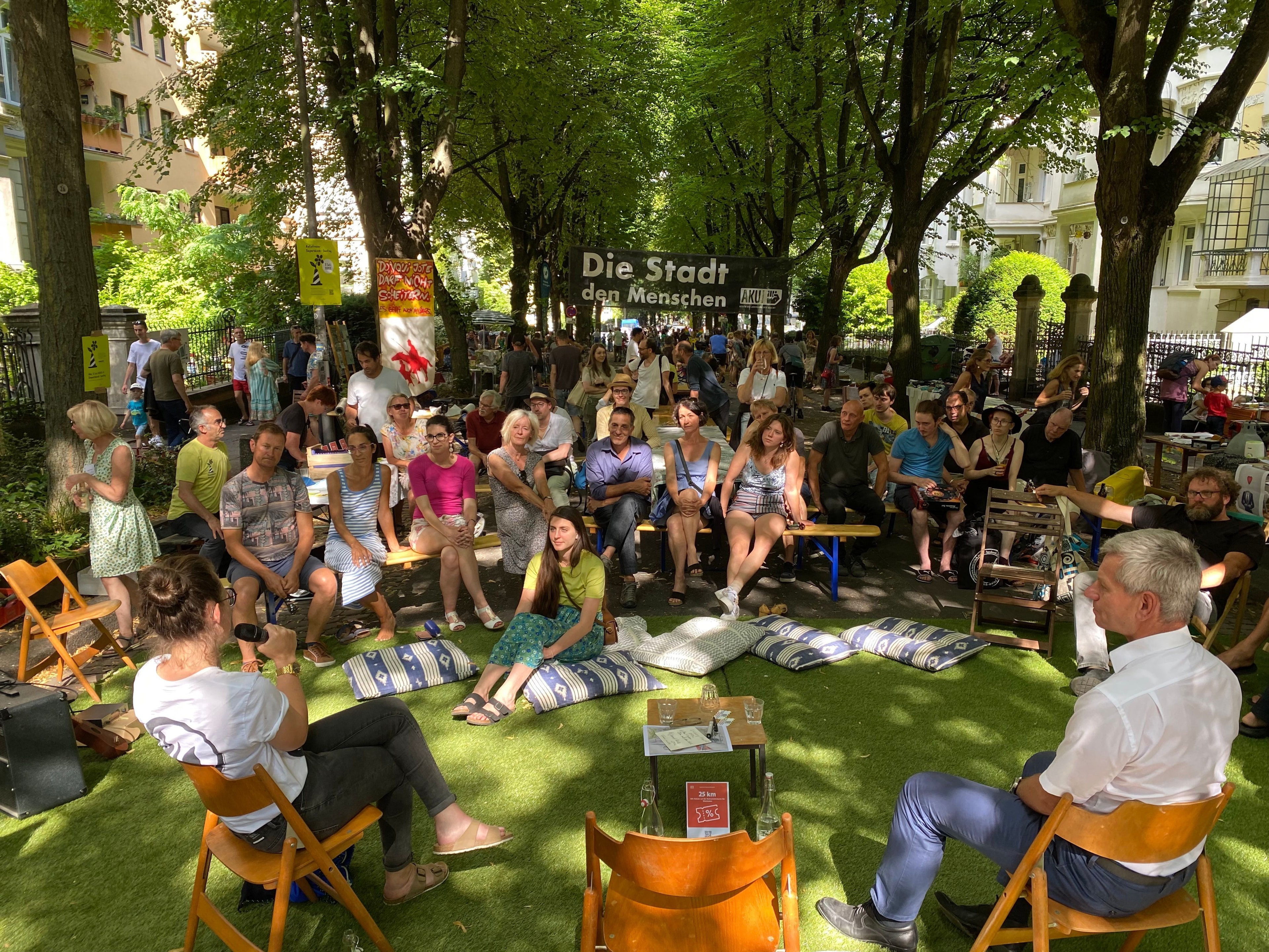 Einige Menschen haben sich auf einer Straße versammelt und hören einem Vortrag zu.