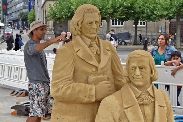 Ein Mann formt Sandskulpturen und wird dabei von einigen Leuten beobachtet.