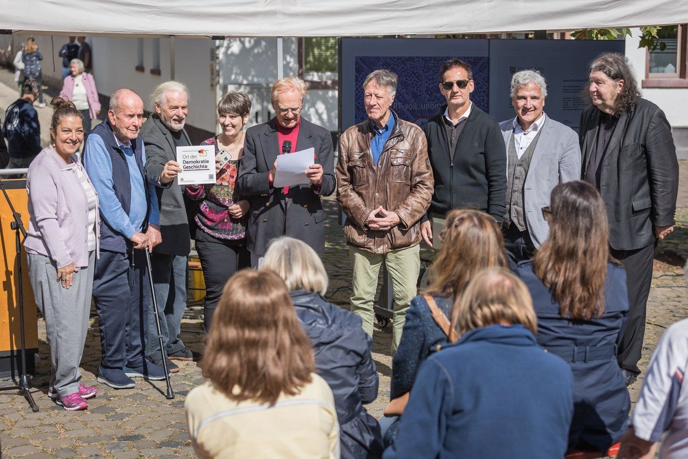 Auf einem Platz stehen draußen ein paar Menschen vor Publikum aufgereiht. Einer von ihnen sagt etwas in ein Mikrofon.