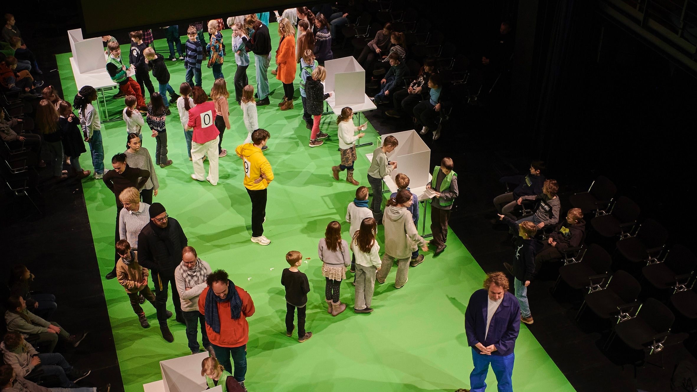 Viele Kinder und Erwachsene simulieren auf einer Bühne eine Wahlkabine