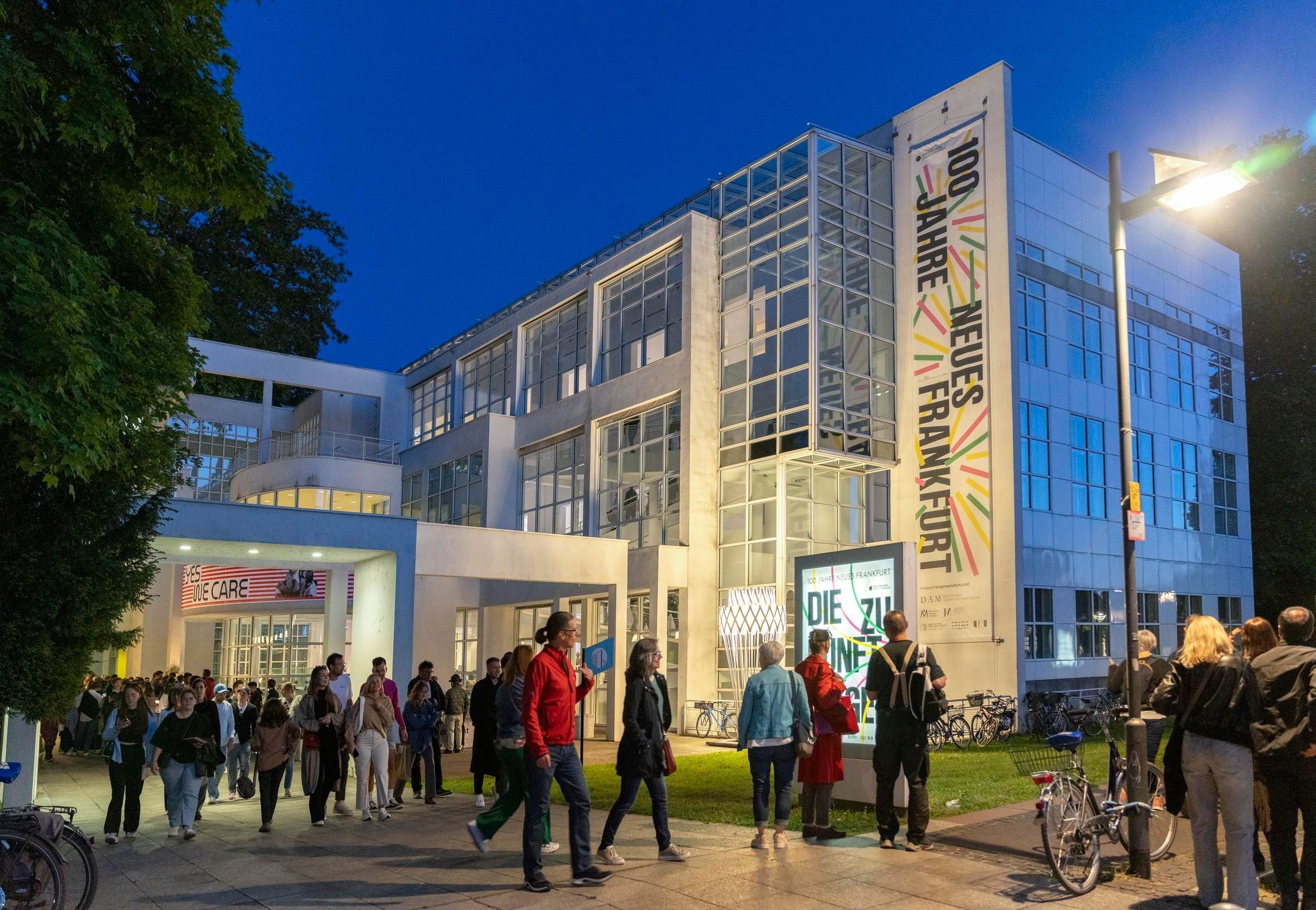 Menschen laufen vor dem Museum Angewandte Kunst in Frankfurt. Im Hintergrund sieht man die aktuellen Ausstellungen 100 Jahre Neues Frankfurt.