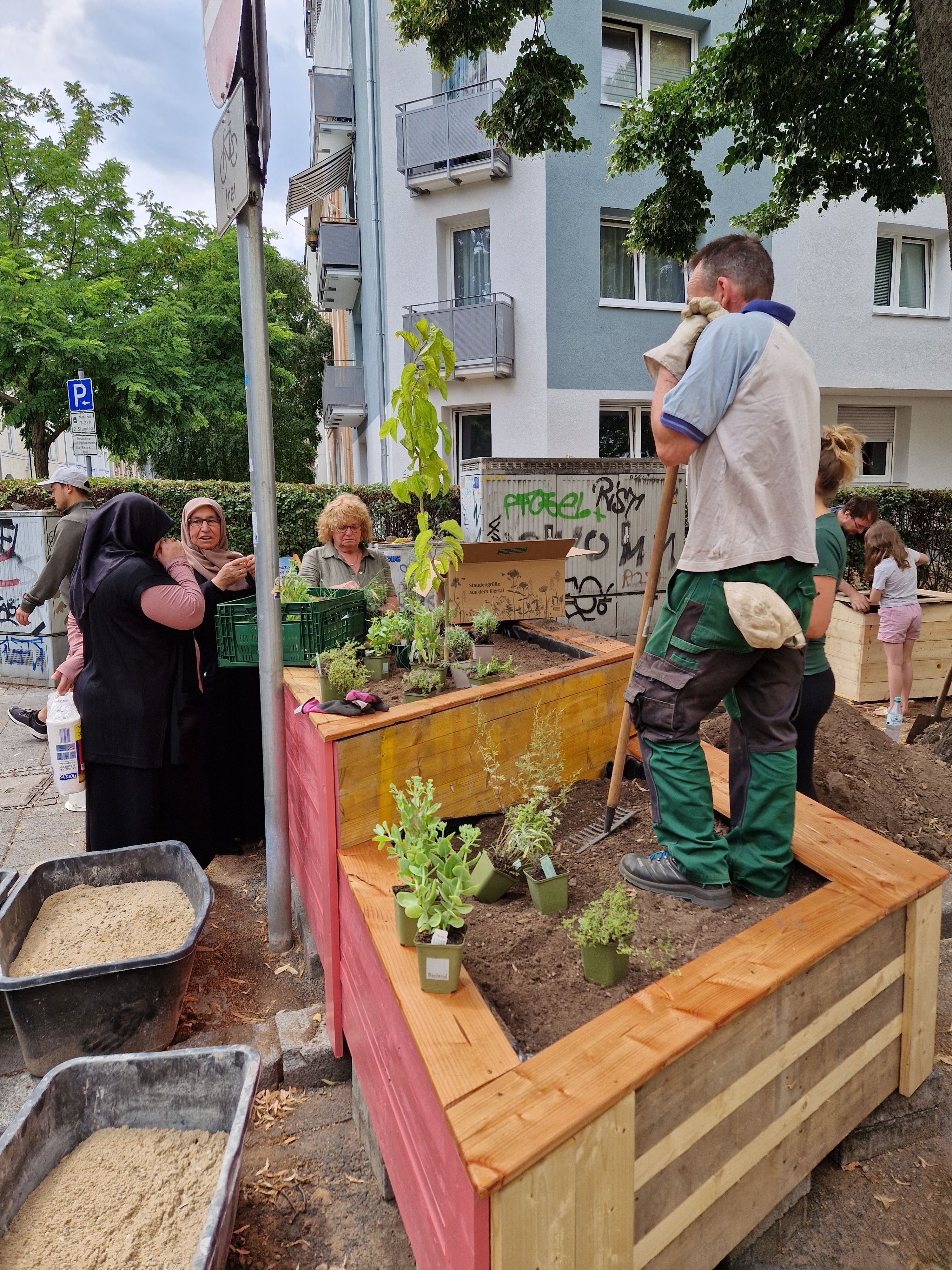 Auf einer Straße stehen Beete. Um und auf ihnen stehen verschiedene Personen.