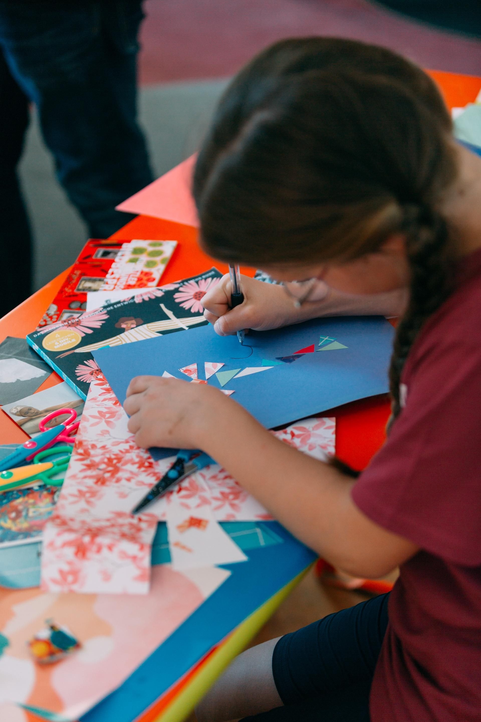 Ein Kind sitzt an einem Basteltisch mit diversen Papieren und schreibt oder malt etwas.