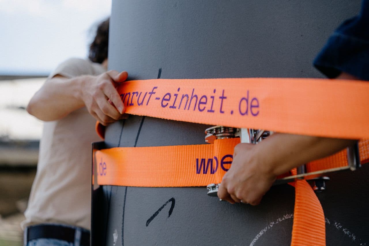 Hände halten einen orangenen Spanngurt, der um eine Säule gespannt ist, fest.