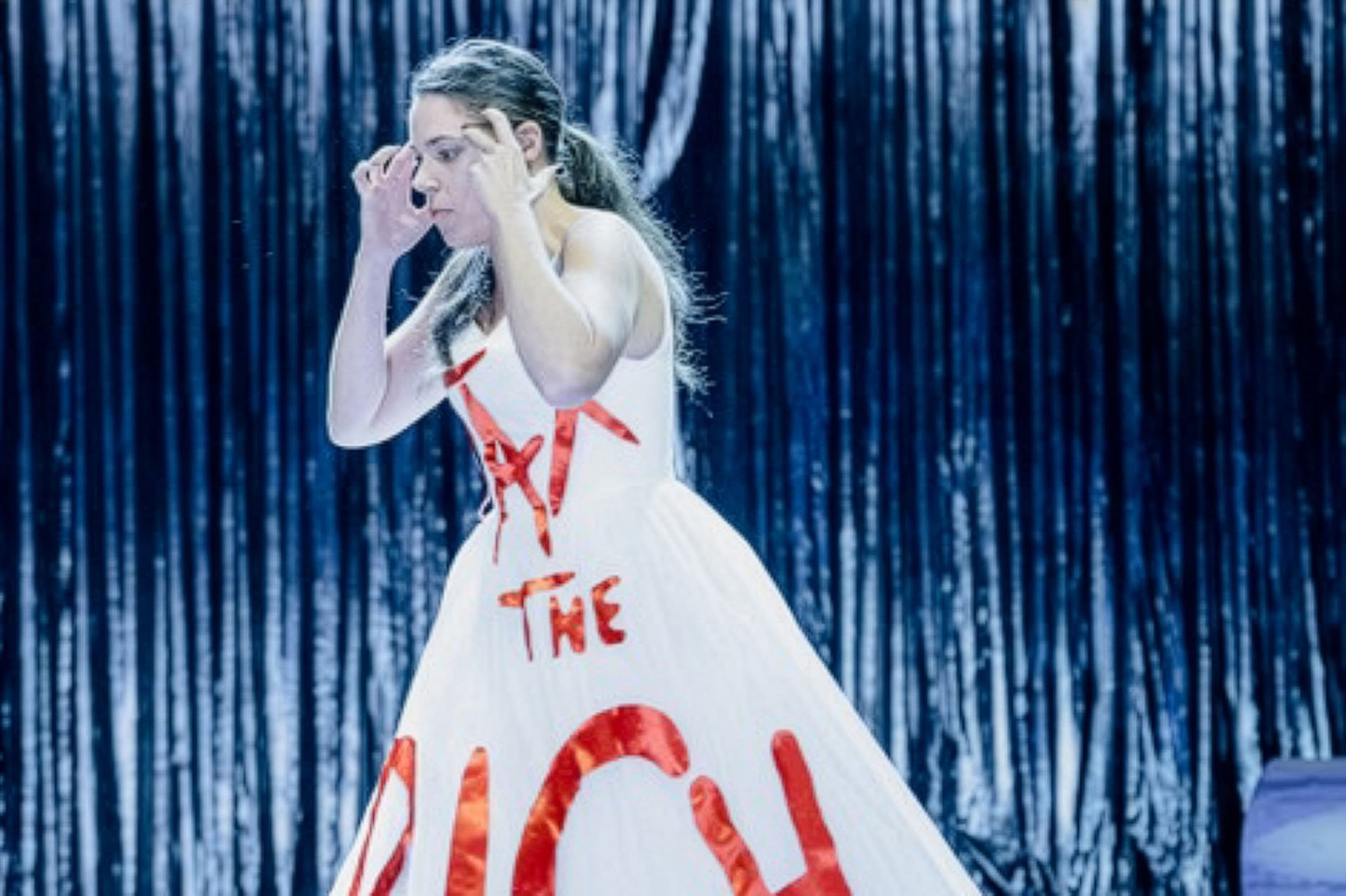 Eine Frau steht auf einer Theaterbühne und trägt ein Kleid mit der Aufschrift "Tax the rich"