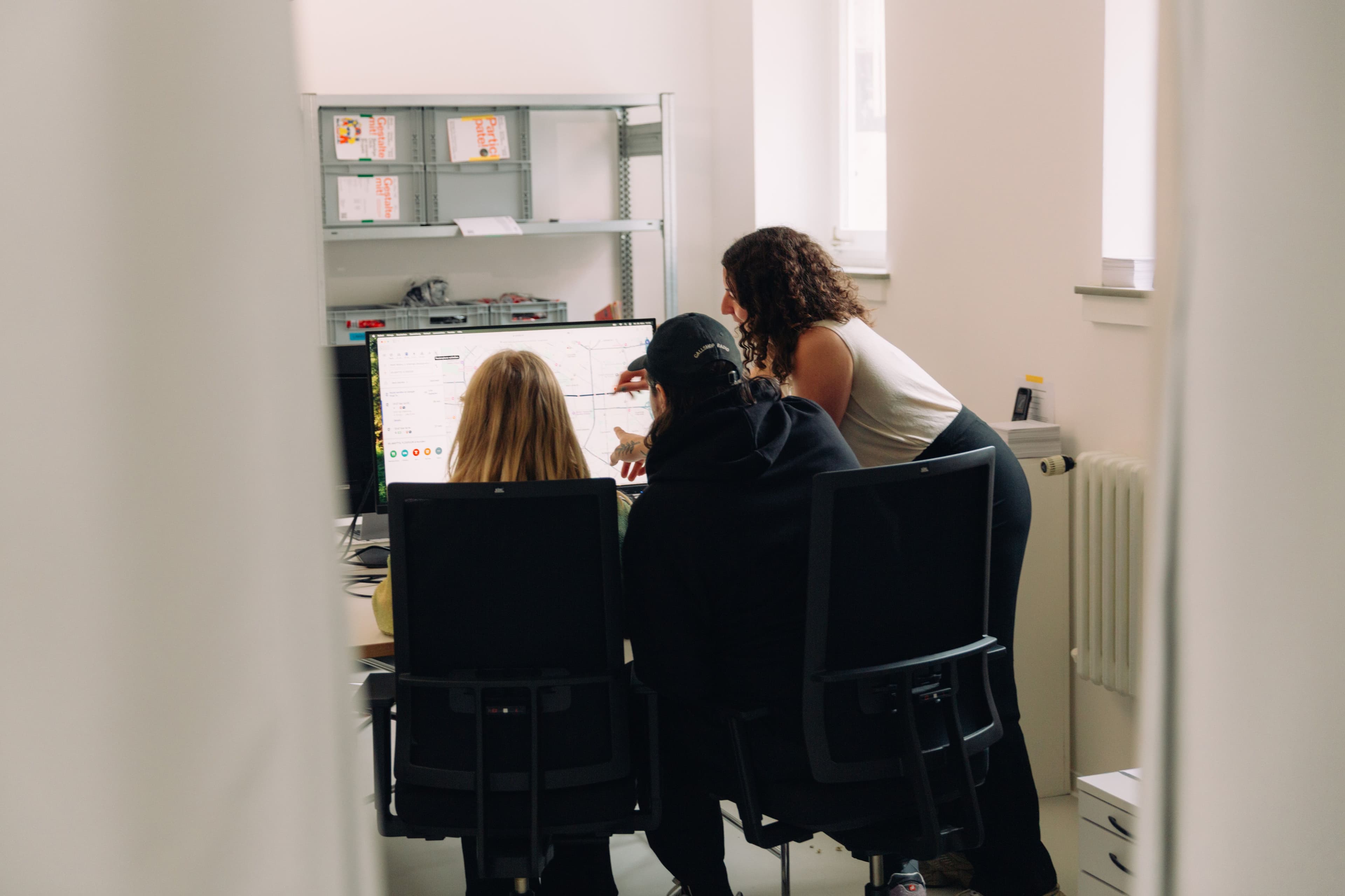 Drei Personen sitzen und stehen an einem Computer-Bildschirm und arbeiten gemeinsam.